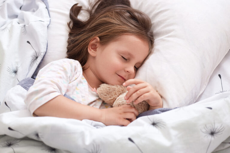 Close up portrait of beautiful girl sleeping in pajama in bed with her teddy bear, lying on pillow with closed eyes, charming cute female kid having dark hair. Childhood and morning time concept.